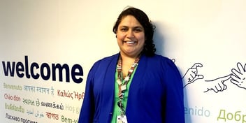 A woman stands smiling in front of a white wall that says “welcome” in many languages including Auslan. She has brown skin and black curly hair. She is dopamine dressing: wearing an electric blue jacket over a grass green linen dress. Her lanyard has a first nations design and has badges saying things like  “she/her” and “proudly neurosparkly”