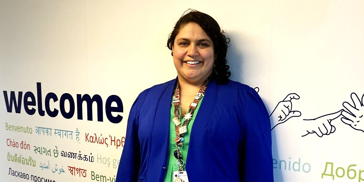 A woman stands smiling in front of a white wall that says “welcome” in many languages including Auslan. She has brown skin and black curly hair. She is dopamine dressing: wearing an electric blue jacket over a grass green linen dress. Her lanyard has a first nations design and has badges saying things like  “she/her” and “proudly neurosparkly”